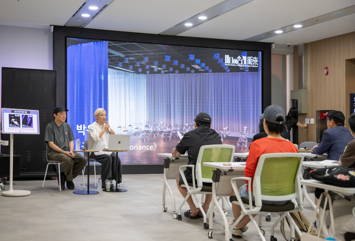 2. 아티스트 토크 : 술래들의 대화, 사진 설명: 강의실에서 발표자 두 명이 대형 화면 앞에 앉아 있으며, 한 발표자가 마이크를 들고 참석자들에게 설명하고 있다. 화면에는 투명한 구형 오브제가 설치된 전시 공간이 보인다.
